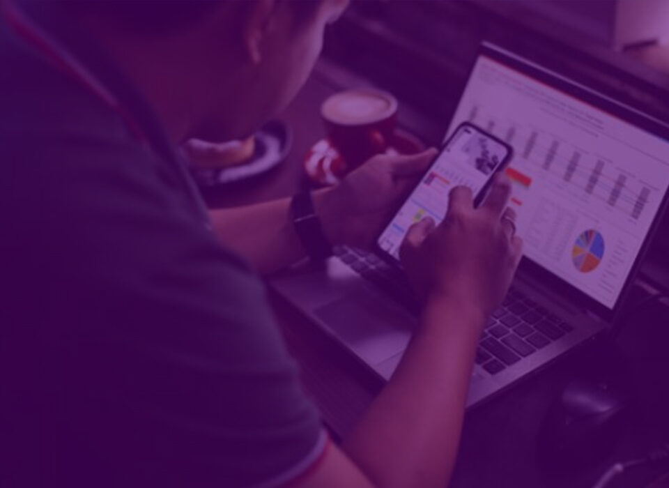 Profissional analisando métricas de tráfego pago em laptop e smartphone, com gráficos de performance e dados de campanhas digitais.